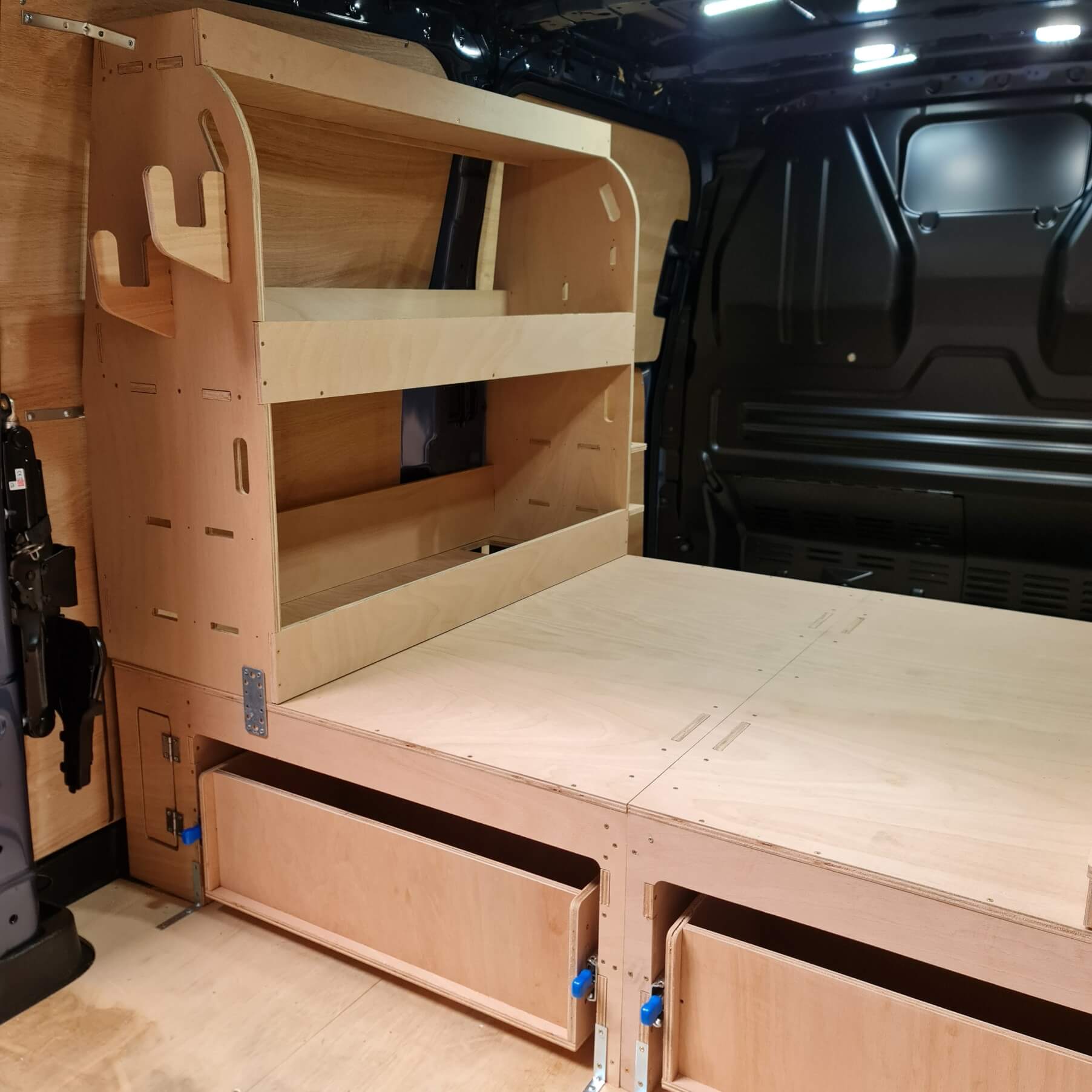 Wooden bed setup inside a vehicle with shelves and drawers.