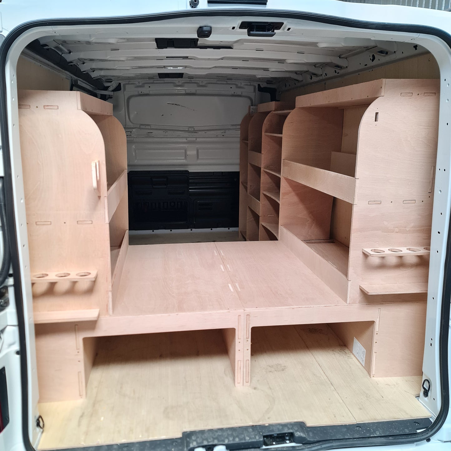 Interior view of a van's cargo area showing a wooden raised floor and storage shelves, with an open door and a viewing window.